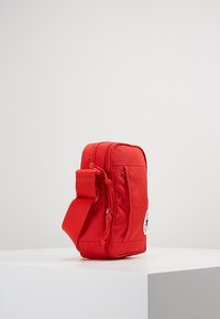 Malá obdélníková červená crossbody taška se dvěma zipovými přihrádky a nastavitelným popruhem, vystavená na bílém podstavci na jednobarevném pozadí.