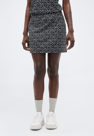 Γυναίκα που φοράει μαύρη φούστα με σχέδια, γκρι κάλτσες και λευκά αθλητικά παπούτσια, στέκεται με τα χέρια χαλαρά στα πλευρά σε λευκό φόντο.