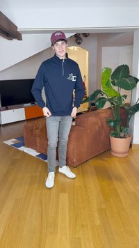 Σκούρος μπλε ζακέτα με φερμουάρ και λευκό λογότυπο, γκρι στενά παντελόνια τζιν και λευκά αθλητικά παπούτσια. Εσωτερικός χώρος με καφέ καπιτονέ καναπέ και γλάστρα με φυτό.