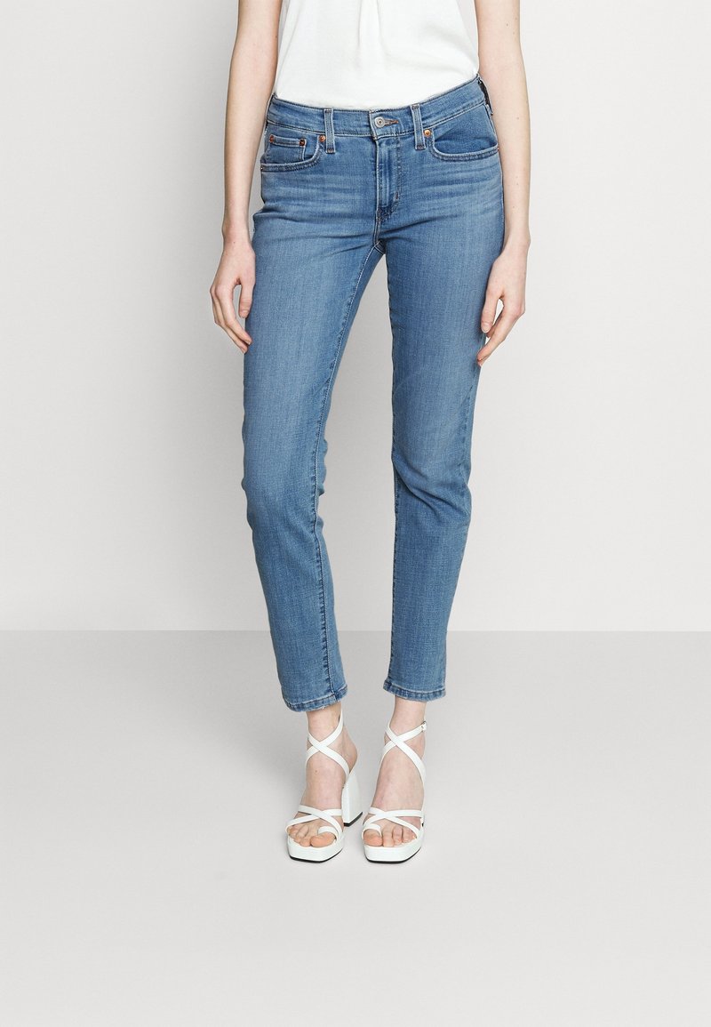 Femme portant un jean slim bleu et des sandales blanches à talons hauts, debout devant un fond uni gris clair.