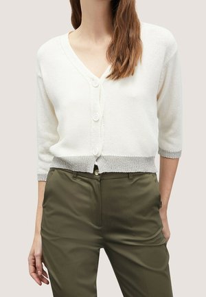 Femme portant un cardigan en maille blanche boutonné avec une bordure grise et un pantalon taille haute vert olive sur un fond uni.