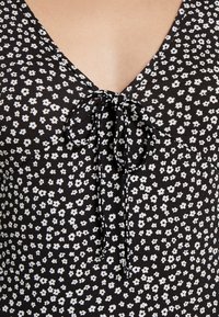 Czarna bluzka z tkaniny z białym kwiatowym wzorem, dekoltem w serek i wiązaniem z przodu. Prążkowana tekstura kontrastuje z gładkimi obszarami.