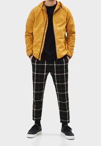 Žlutá lehká bunda, černá mikina a černobílé kárované kalhoty. Černé tenisky s bílými podrážkami doplňují outfit.