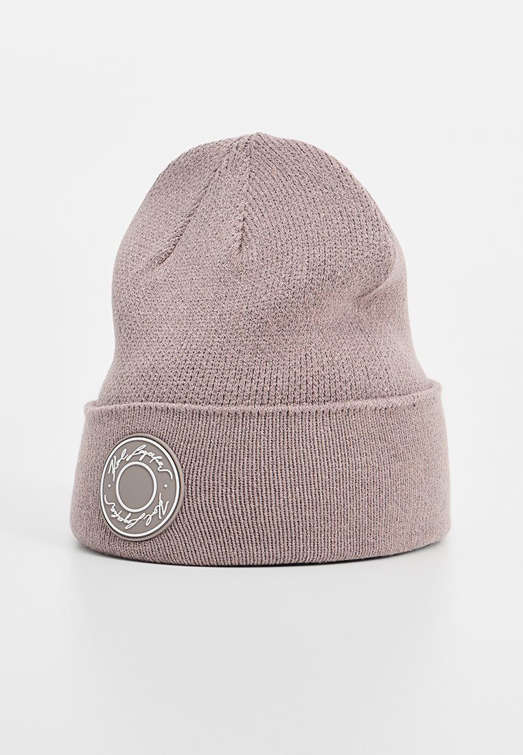 Gebreide beanie in licht mauve, met een opgevouwen cuff en een ronde patch met tekst en embleem, gemaakt van zacht, gestructureerd materiaal.