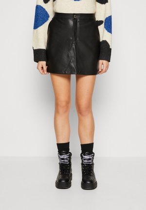 Femme portant une minijupe en cuir noire, des bottes montantes noires avec des lacets blancs, et un pull crème avec des taches bleues et noires.