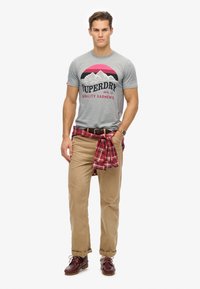 Gray T-shirt with printed mountains and text, tan chinos, red and black checkered flannel tied at the waist, brown shoes.