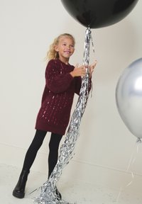 Vijoličen pleten pulover z bleščicami, črne gamaše in črni gležnjarji. Drži črno balon z srebrnimi trakovi ob srebrnem balonu.