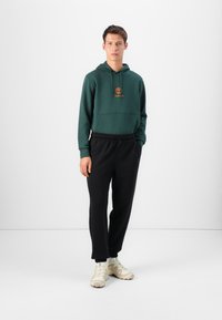 Grønn bomull hettegenser med frontlomme og oransje logo, kombinert med svarte joggebukser med elastisk midje og beige treningssko.