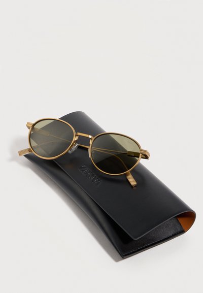 Okrúhle slnečné okuliare so zlatým rámom a zelenými šošovkami, uložené na zatvorenom čiernom koženom púzdre s vyrazeným nápisom "ZARA".
