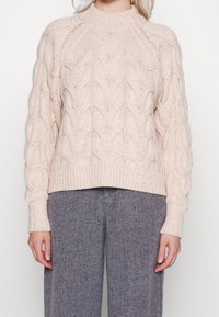 Femme portant un pull en maille épaisse couleur crème avec des motifs en losange et un pantalon gris en chevrons, se tenant devant un fond uni.