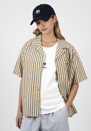 Kvinde iført en navy kasket, beige og gråstribet kortærmet skjorte over en hvid top med broderi og lysegrå bukser med den ene hånd i lommen.