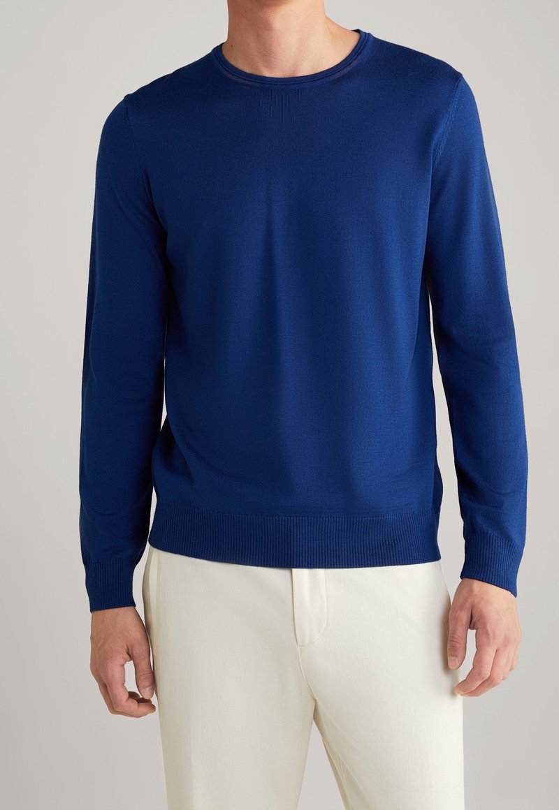 Homme portant un pull à col rond bleu uni à manches longues et un pantalon beige clair, debout devant un arrière-plan neutre.