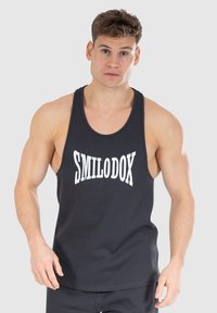 Muskularny mężczyzna w czarnej koszulce na ramiączkach z białym napisem "SMILODOX", stojący na jednolitym jasnoszarym tle.