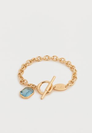 Gouden kettingarmband met een toggle-sluiting met een charm van een blauwe edelsteen en een klein gegraveerd ovaal label.