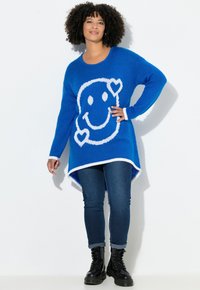 Blauwe gebreide trui met wit smiley gezicht ontwerp, geribbelde manchetten en een hoge-lage zoom. Gecombineerd met donkere jeans en zwarte veterlaarzen.