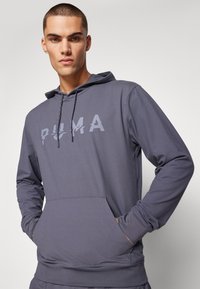 Grauer Kapuzenpulli mit Kängurutasche, ausgestattet mit einem großen "PUMA"-Logo in einer helleren Farbe. Glatte Textur und verstellbare Kordeln.