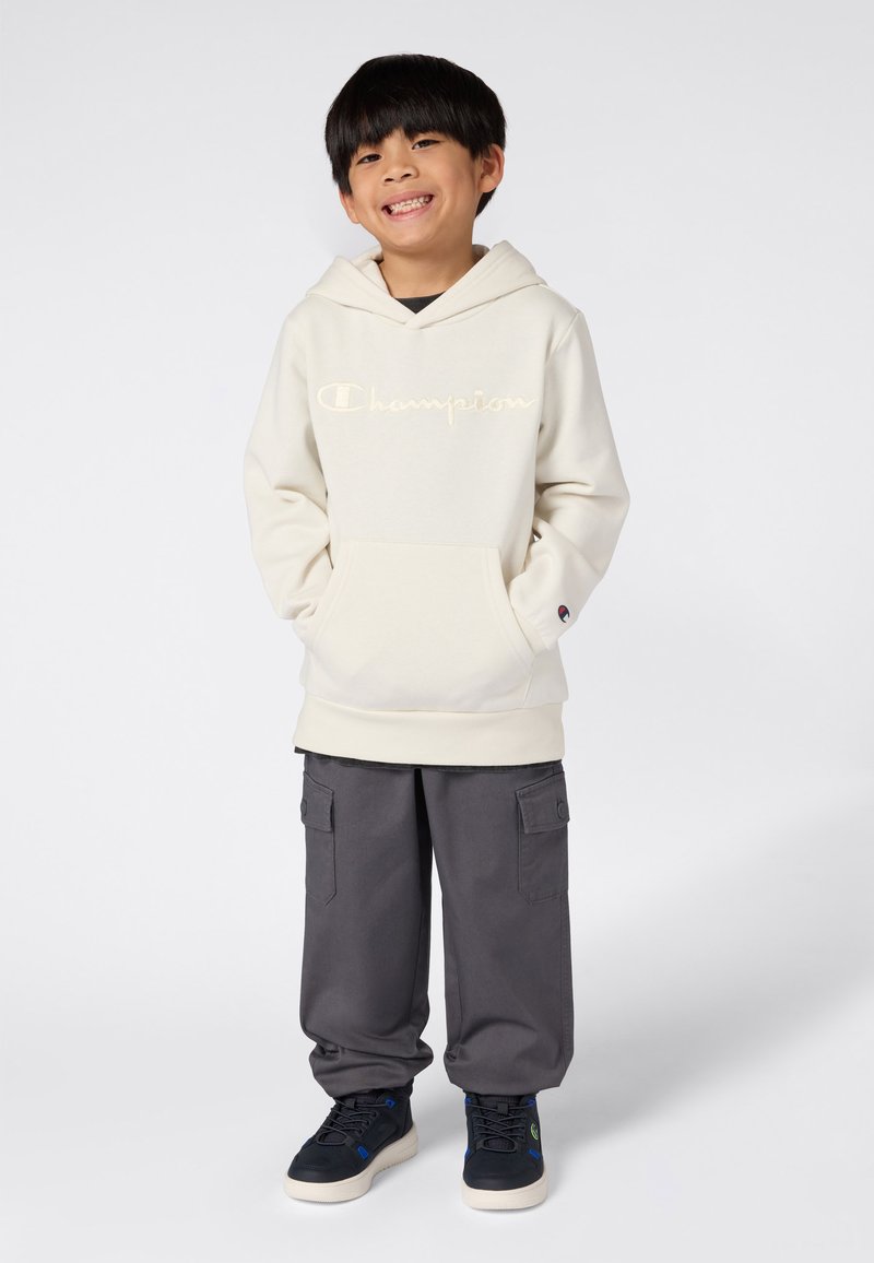 Niño pequeño sonriendo con las manos en los bolsillos, vistiendo una sudadera de crema de Champion, pantalones cargo grises y zapatillas negras sobre un fondo blanco.