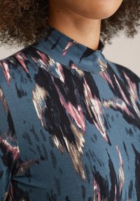 Pull col roulé multicolore en tissu doux, avec un fond bleu et des motifs abstraits dans des tons rose, noir et beige.