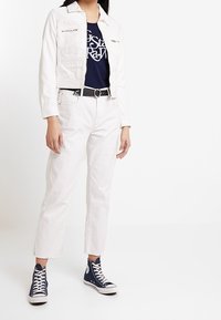 Bílá zkrácená džínsová bunda s kapsami na prsou, námořnické tričko s grafikou, bílé džíny rovného střihu s roztrhanými manžetami a modré vysoké tenisky.