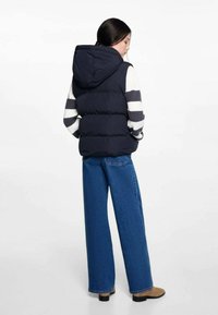 Mörkblå pufferväst med huva, med en kviltad design. Modellen bär en randig tröja och vida blå jeans med tan-färgade stövlar.