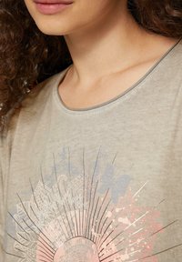 Helle beige T-Shirt mit Rundhalsausschnitt, das ein gedrucktes Design in Pastellfarben zeigt, einschließlich Schmetterlingen und komplizierten Mustern.
