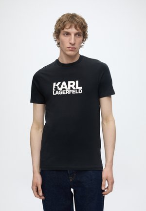 Młody mężczyzna o jasnobrązowych włosach, ubrany w czarną koszulkę Karl Lagerfeld i ciemnoniebieskie jeansy, stoi na jednolitym, jasnym tle.