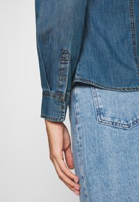 Denimskjorta med en blå toning, med knäppning vid ärmslut och sömdetaljer. Tyget är texturerat med en slät yta och en avslappnad passform.
