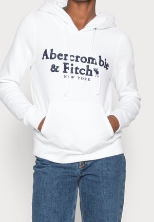 Henkilö pukeutuneena valkoiseen Abercrombie & Fitch -huppariin, jossa kengurutasku, yhdistettynä sinisiin farkkuihin, yksinkertaisella taustalla.