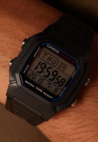 Montre numérique noire avec un cadran carré, accents bleus et rouges, étanche jusqu'à 100 m, affichant le jour et l'heure. Bracelet en caoutchouc.