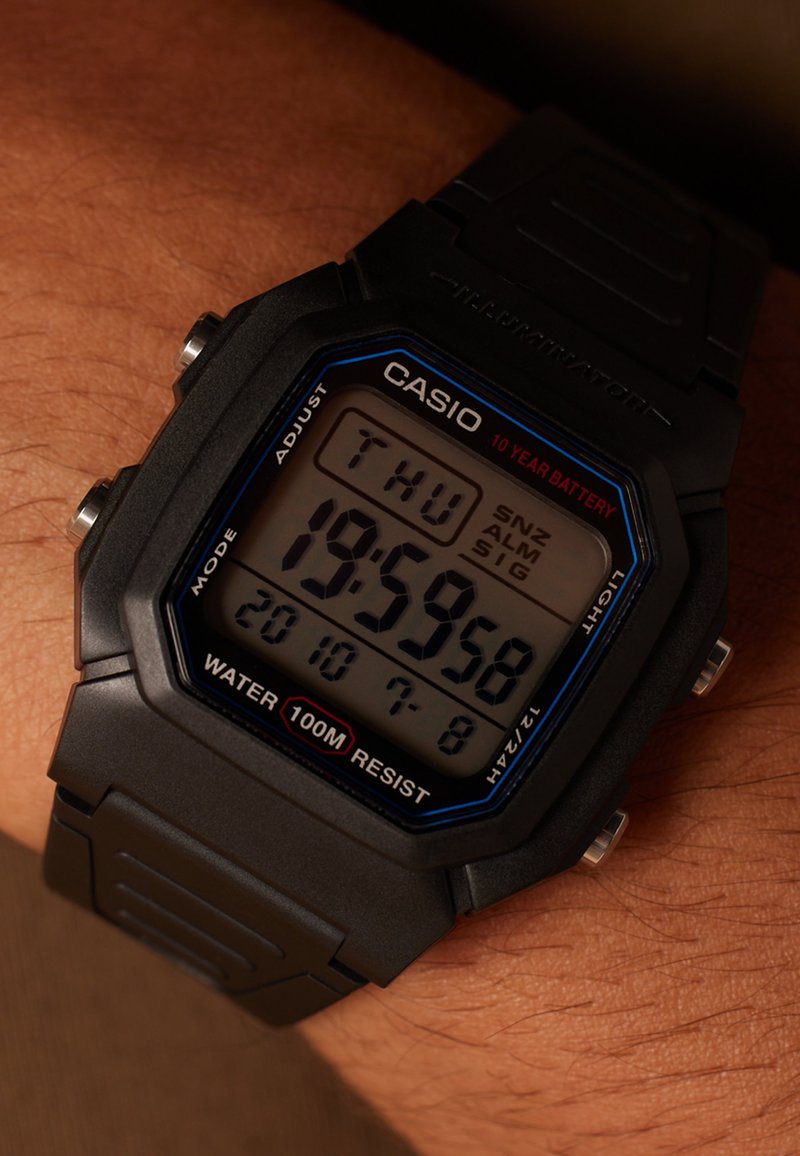 Montre numérique noire avec un cadran carré, accents bleus et rouges, étanche jusqu'à 100 m, affichant le jour et l'heure. Bracelet en caoutchouc.
