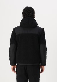 Sort jakke med en blanding af fuzzy fleece og glat stof, der har en hætte, elastisk kant og kontrasterende paneldetaljer på skuldrene.