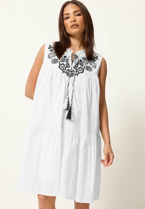 Femme portant une robe blanche sans manches avec broderie florale noire et liens à pompons au niveau du col, debout devant un fond uni.