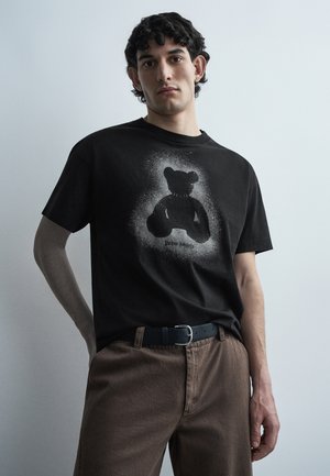 Νέος άνδρας με σγουρά σκοτεινά μαλλιά στέκεται μπροστά σε μονόχρωμο φόντο φορώντας μαύρο μπλουζάκι με εικόνα αρκουδάκι και καφέ παντελόνι με μαύρη ζώνη.