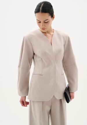 Beigefarbener Blazer mit tiefem V-Ausschnitt, strukturierten Schultern und seitlichen Taschen. Glattes Material; kombiniert mit passender Hose und einer schwarzen Clutch.