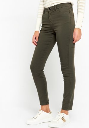 Personne portant un pantalon slim vert olive foncé, des baskets blanches avec des rayures dorées, et un haut blanc à manches longues avec des boutons dorés sur les manches.