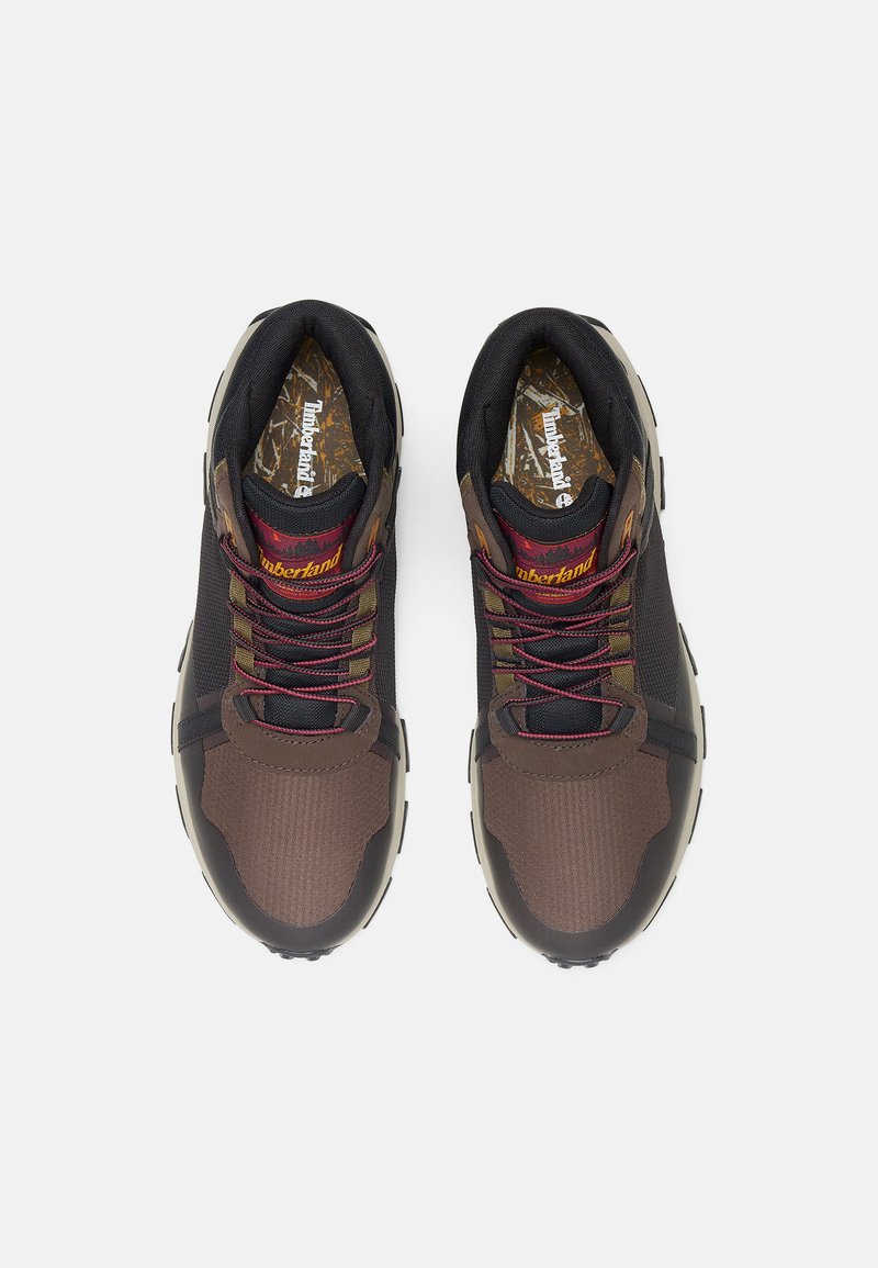 Bruine en zwarte sportschoenen met een stoffen bovenkant, textuurpanelen, contrasterende rode veters en rubberen buitenzool. Timberland-logo op de binnenzool.