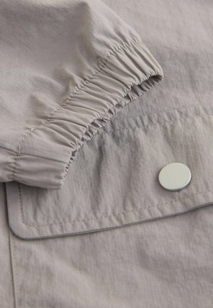 Hellgrauer Stoff mit leichtem Glanz, ausgestattet mit einem gerafften elastischen Bündchen und einem silbernen Druckknopf an einer Taschenklappe. Glatte Textur und minimalistisches Design.