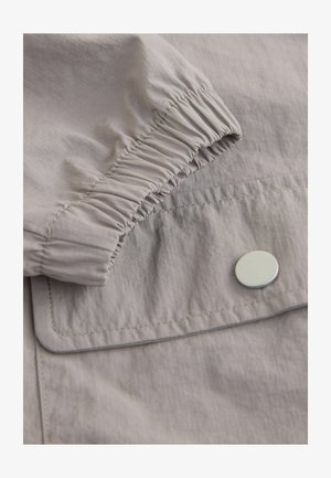 Light gray fabric with slight sheen, featuring gathered elastic cuff and silver snap button on a pocket flap. Smooth texture and minimalist design.