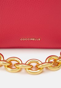 Czerwona, fakturowana skórzana powierzchnia z małym, złotym logo "COCCINELLE" oraz błyszczącym, złotym, masywnym łańcuchem poniżej na białym tle.
