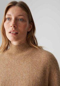 Pull à col roulé en laine beige avec texture côtelée et coupe ample. Gros plan montrant le col et l'aspect doux et chaud.