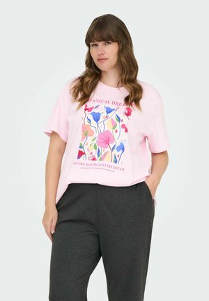 Femme aux longs cheveux ondulés portant un t-shirt graphique fleuri rose et un pantalon gris foncé, se tenant avec une main dans sa poche.