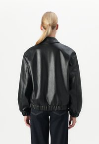 Kvinne med blond hestehale iført en svart skinnjakke med elastiske mansjetter og midjebånd, kombinert med mørkeblå jeans, vendt bort.