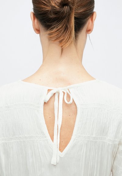 Barna haját lófarokba fogó nő, fehér, texturált blúzt visel, melynek hátulján kulcslyuk alakú nyílás és a nyaknál masni van.