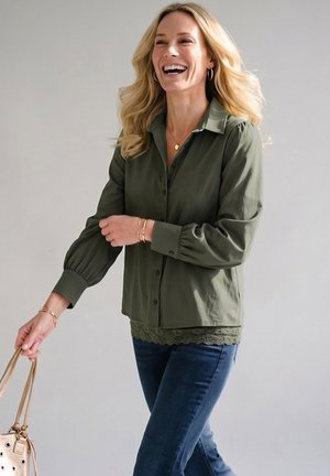 Lachende vrouw met blond haar draagt een olijfgroene blouse, blauwe jeans, gouden sieraden en houdt een beige handtas vast tegen een effen achtergrond.