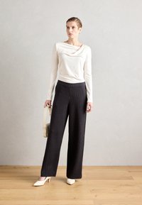 Langermet kremfarget topp med drapert halslinje, paired med høytlivs, vidde svarte stripete bukser. Modellen holder en beige håndveske.