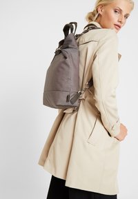Mochila de tela gris con un diseño estructurado, correas ajustables y herrajes metálicos. Presenta una textura suave y un aspecto minimalista.