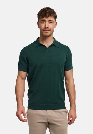 Man met kort krullend haar, gekleed in een donkergroen poloshirt met korte mouwen en beige broek, staande tegen een effen lichte achtergrond.