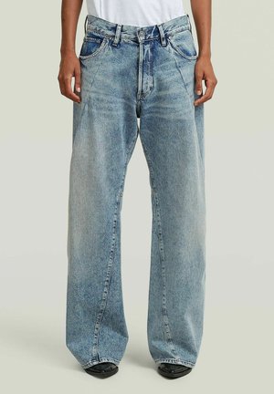 Person trägt locker sitzende, weit geschnittene hellblaue Jeans mit hoher Taille und Fronttaschen, kombiniert mit schwarzen Schuhen und einem weißen Oberteil.