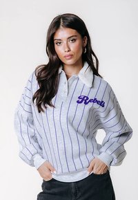 Jovem mulher com longos cabelos escuros, vestindo uma camisa riscada em cinza e roxo com o texto "Rebels", de pé com as mãos nos bolsos, fundo neutro.
