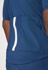 Mørkeblått treningskjorte med stretchy materiale, tettsittende design, hvite striper og en glidelåslomme bak.